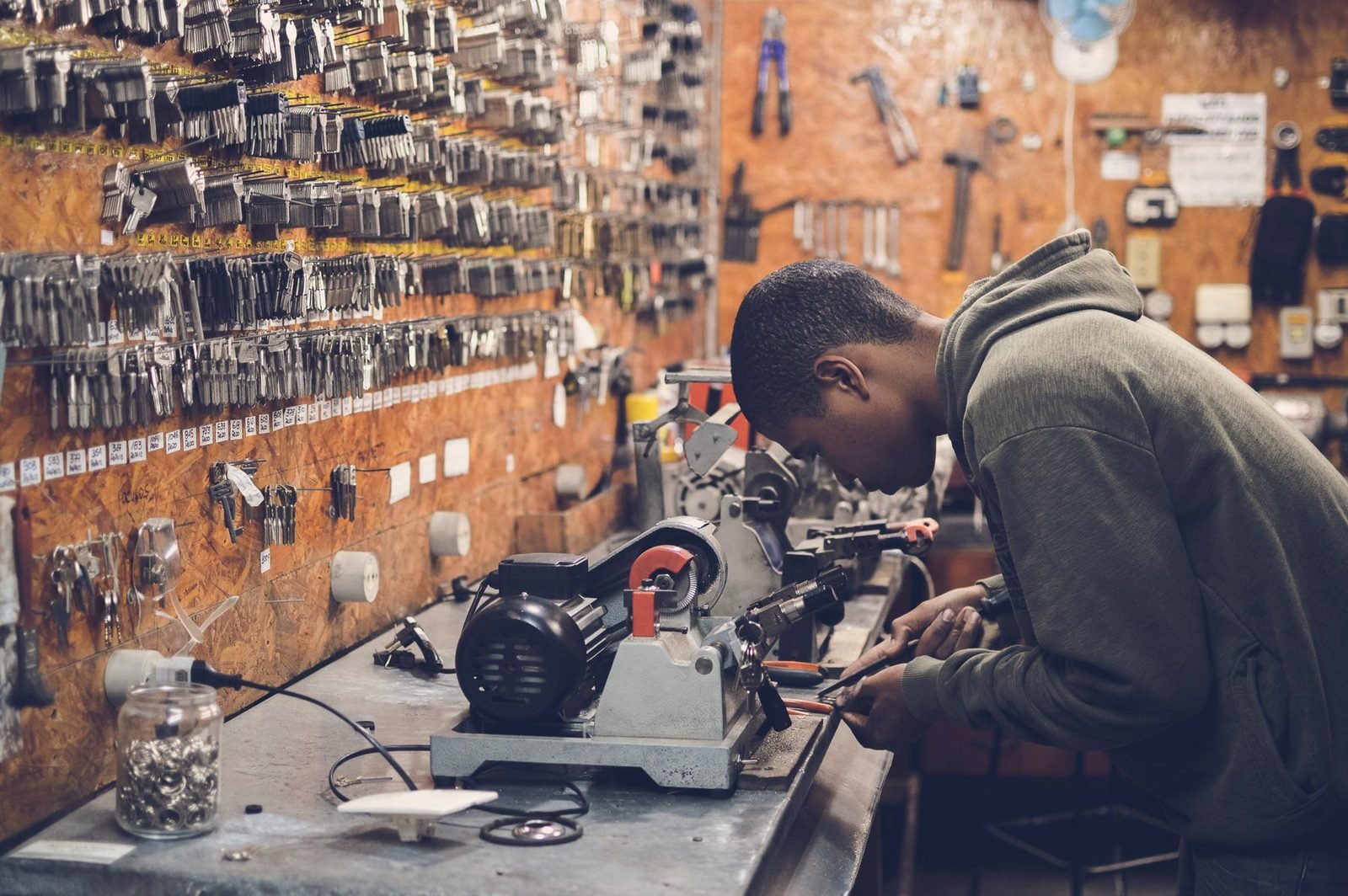 A man bent over a key-cutting machine at a workbench, a wall of key blanks behind him