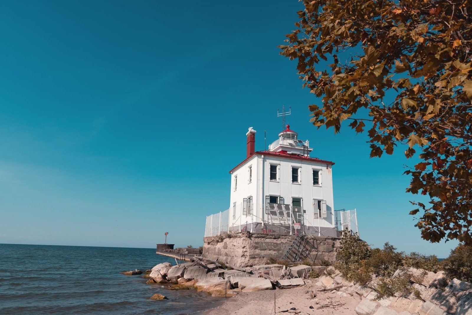 A white lighthouse on a rocky breakwater under a clear blue sky, autumn leaves in the corner